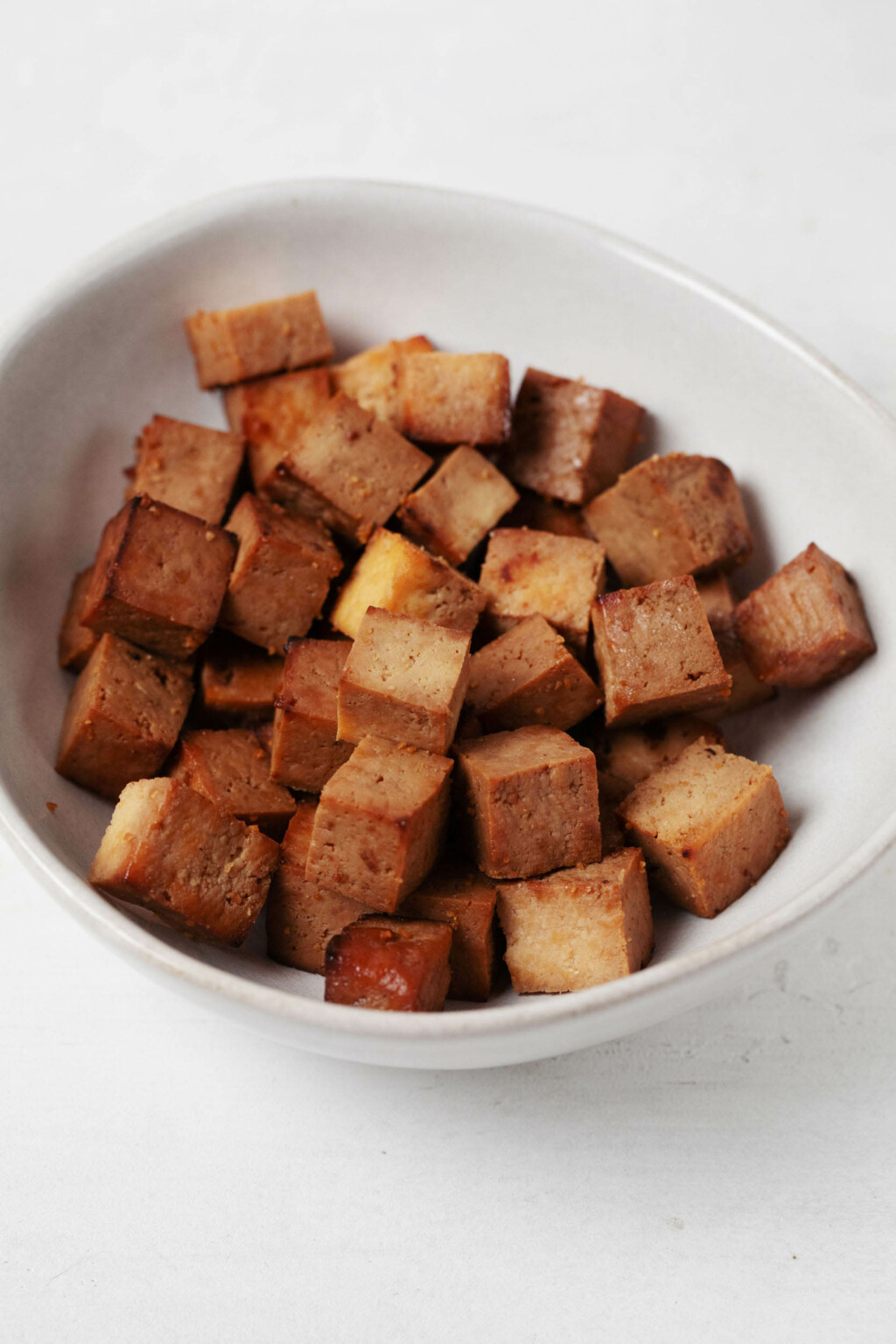 Baked teriyaki tofu cubes rest in a round, asymmetrical white bowl.
