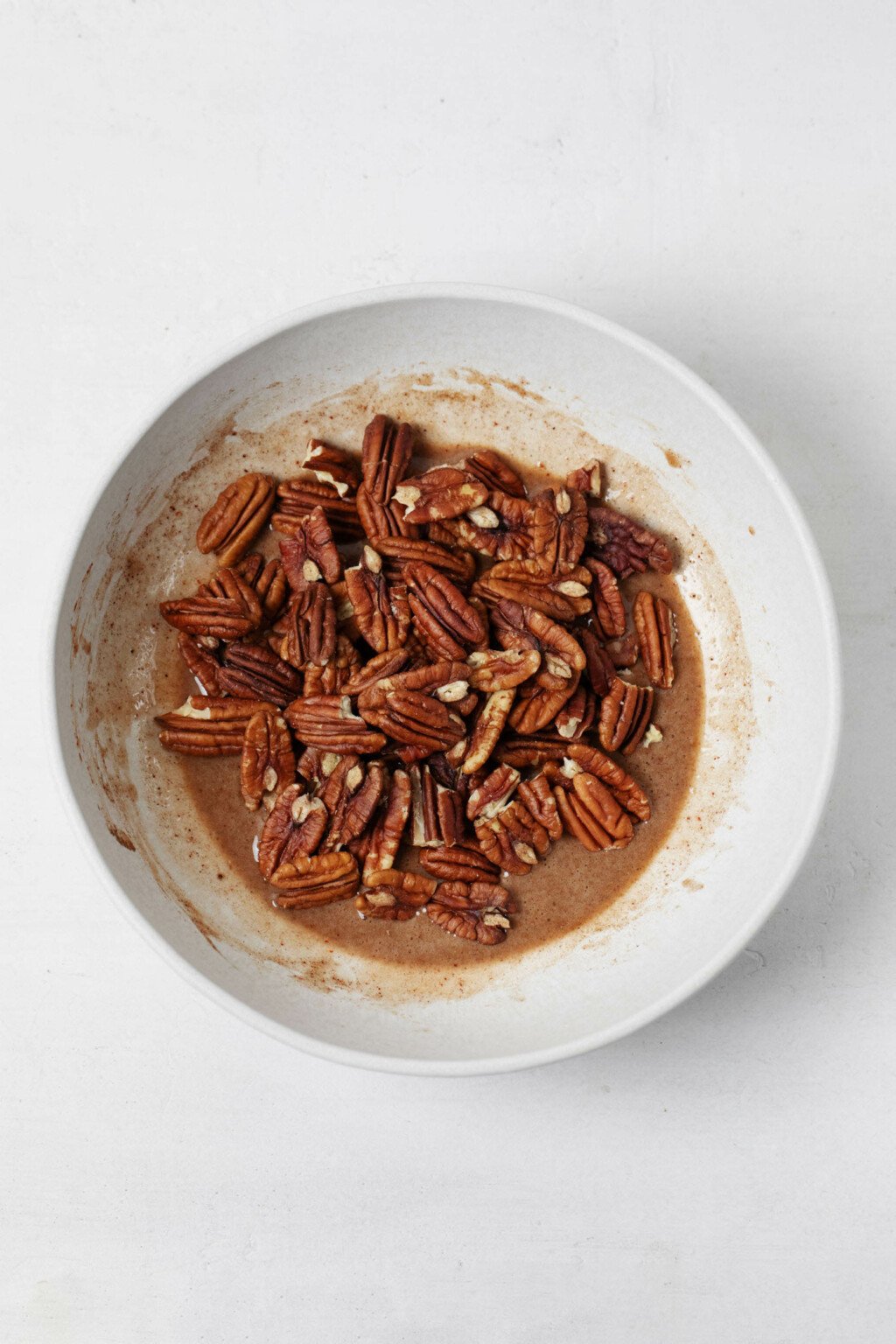 A small white bowl is filled with pecans that are being glazed in a viscous liquid. It rests on a white surface.