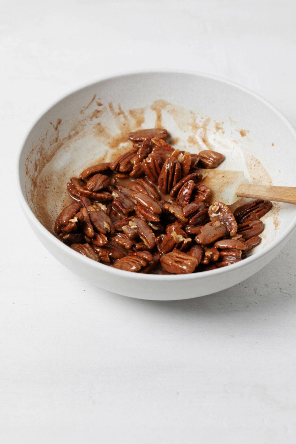 Pecan halves are being mixed with a sugary, liquid coating for baking in the oven. They're in a white mixing bowl that's resting on a white surface.