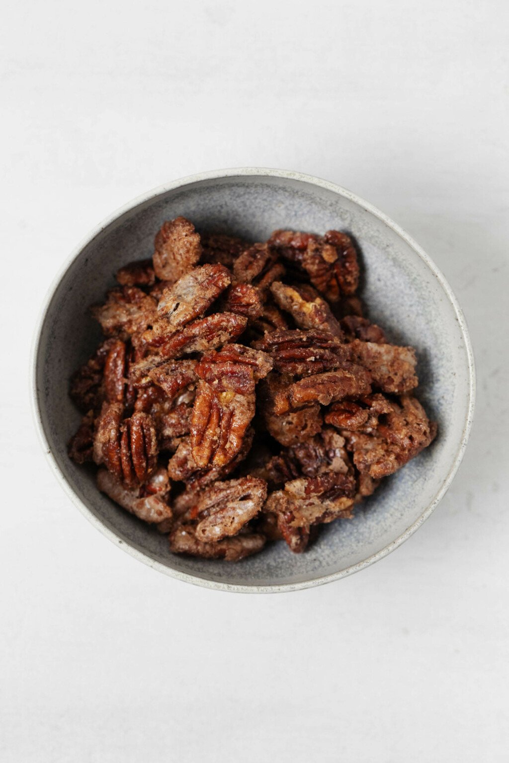 Crispy, vegan candied pecan halves are resting in a gray, ceramic bowl. It's resting on a white suface.