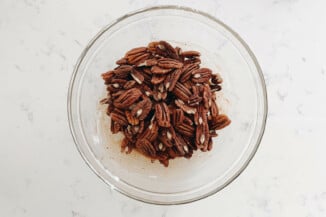 A clear, pyrex bowl is filled with pecans that are being glazed in a viscous liquid. It rests on a white surface.