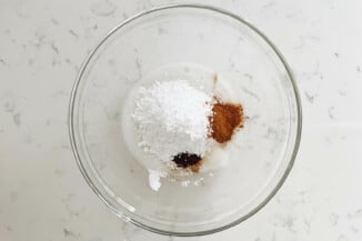 A clear, pyrex bowl is filled with powdered sugar, spices, and a clear liquid. It rests on a white surface.
