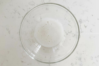 A small, clear pyrex bowl is filled with beaten aquafaba, or chickpea water. It has a frothy appearance.