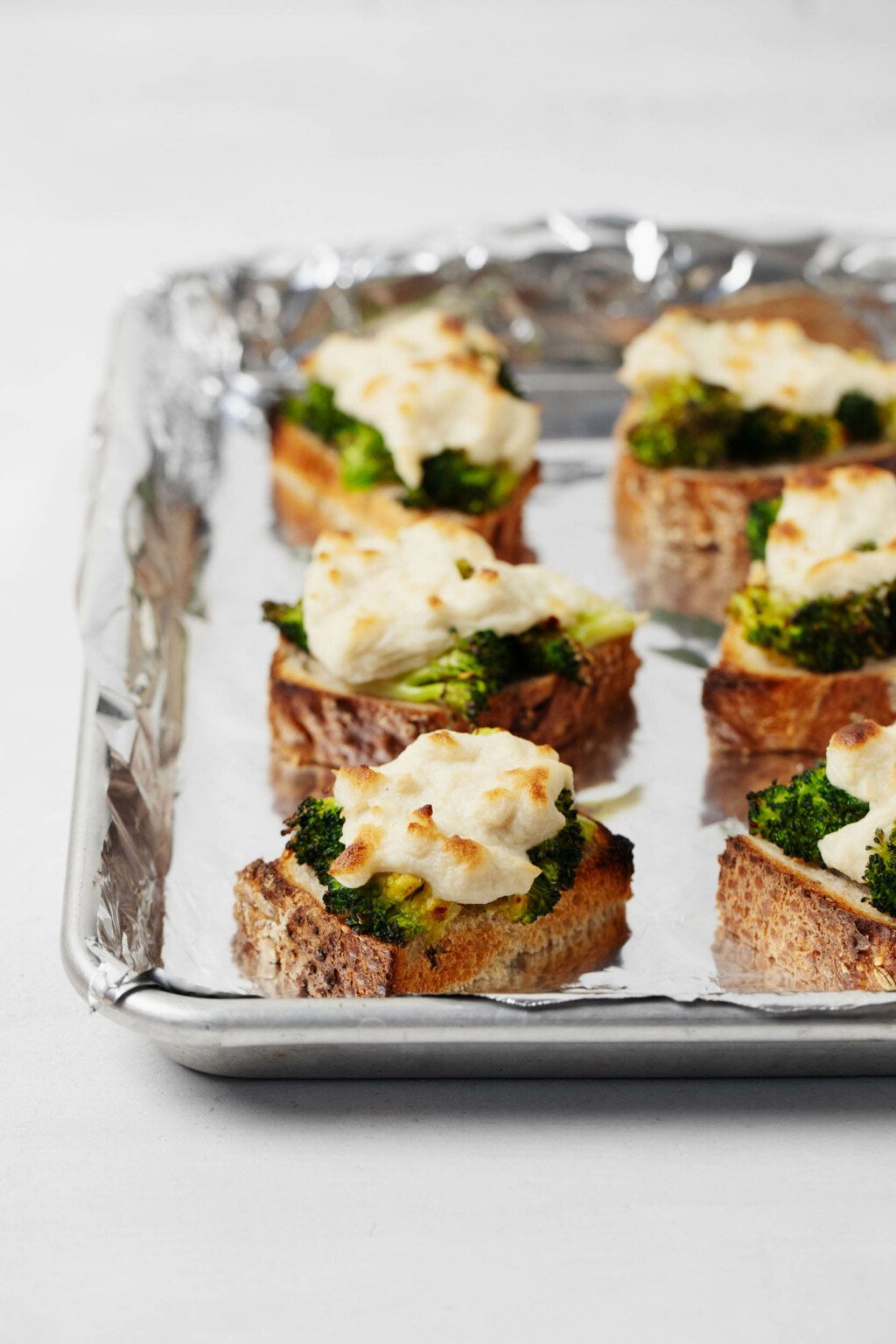 Slices of bread, each topped with cooked broccoli and toasted mozzarella cheese, are lined up on a baking sheet with foil.