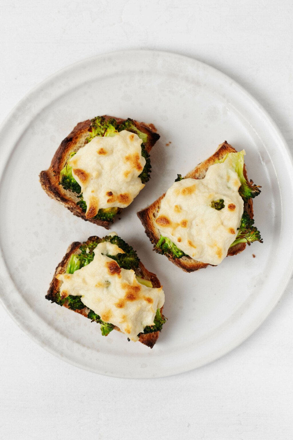 An overhead image of three plant-based broccoli melts on whole grain bread.  They rest on a round, white plate.