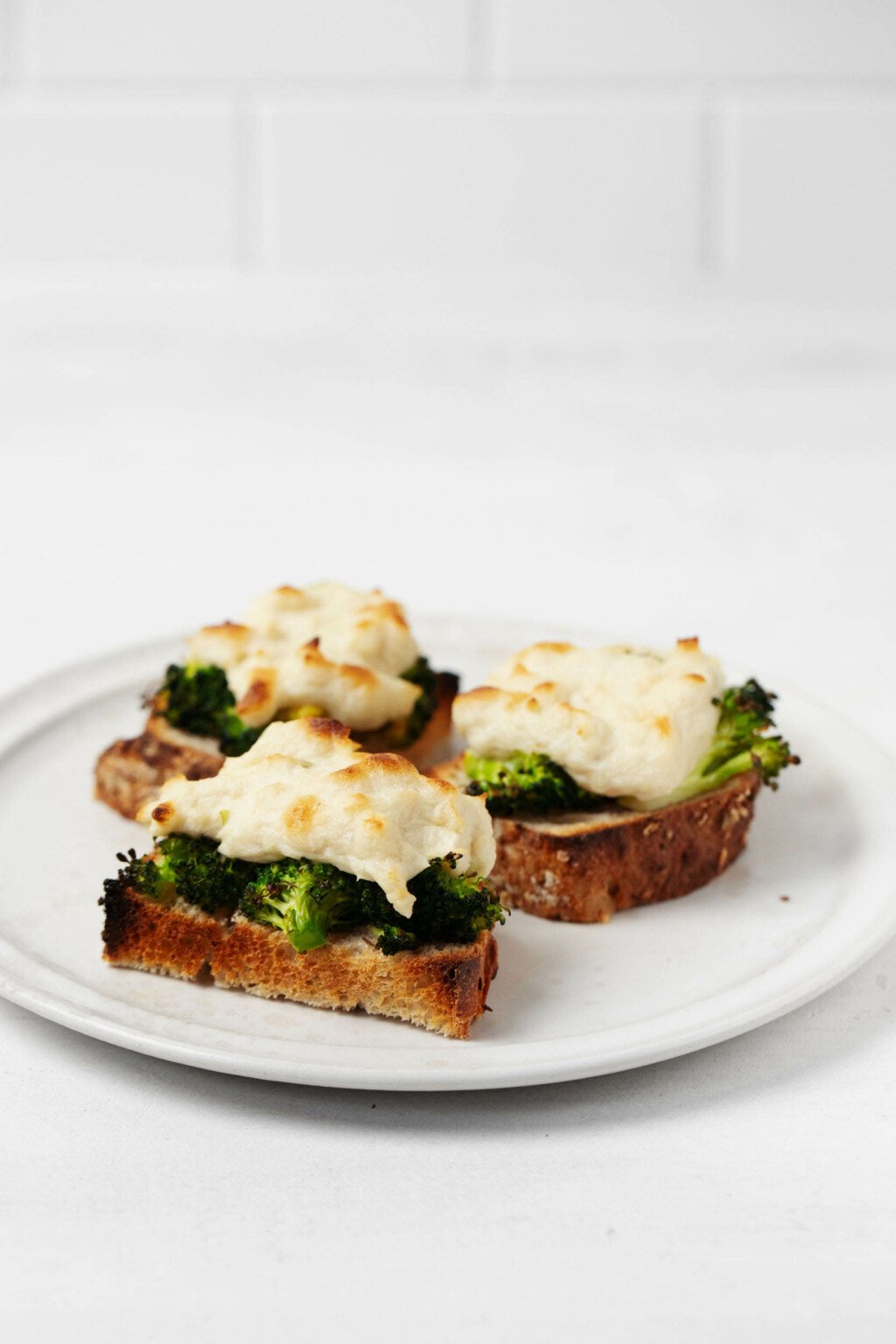 An angled photo of toasted halves of bread, which are topped with a plant-based, mozzarella style cheese and bright green broccoli florets. There's a white tile surface in the background.