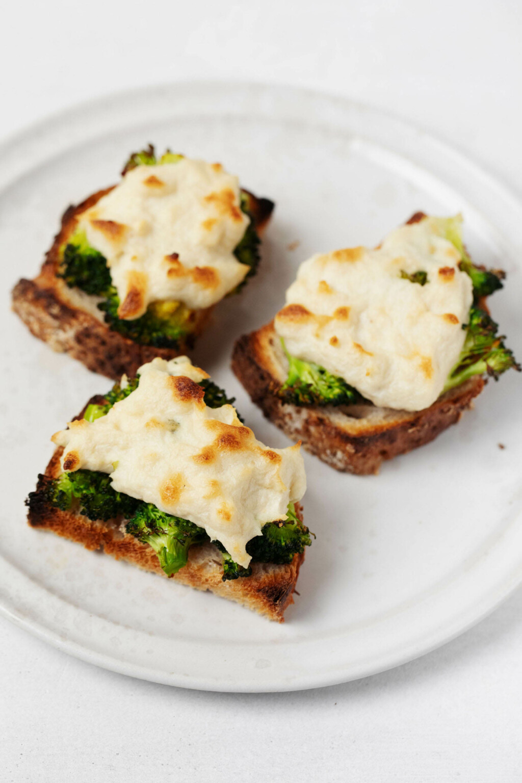 Three half-slices of vegan broccoli melts are laid out on a white, rimmed plate. The plate rests on a white surface.