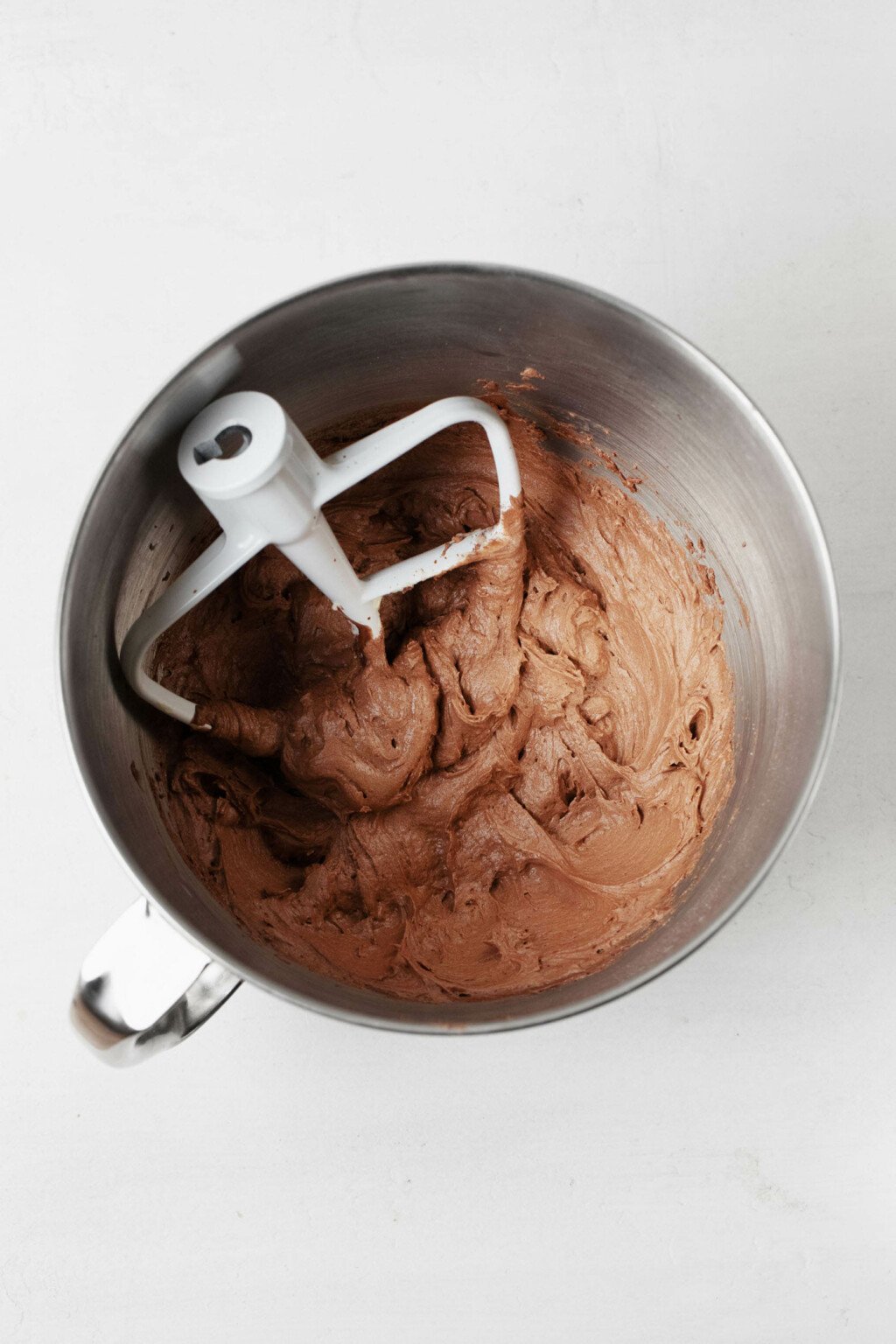 The silver bowl of a stand mixer has just been used to prepare vegan chocolate frosting. The white paddle attachment rests in the bowl.