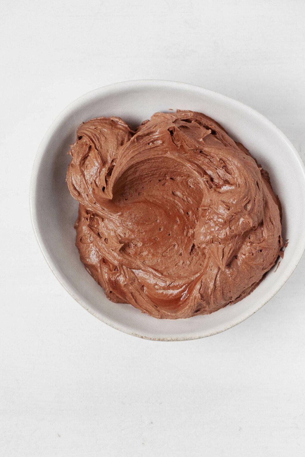 A swirl of vegan chocolate frosting is served in a small, asymmetrical ceramic bowl.  It is located on a white surface.