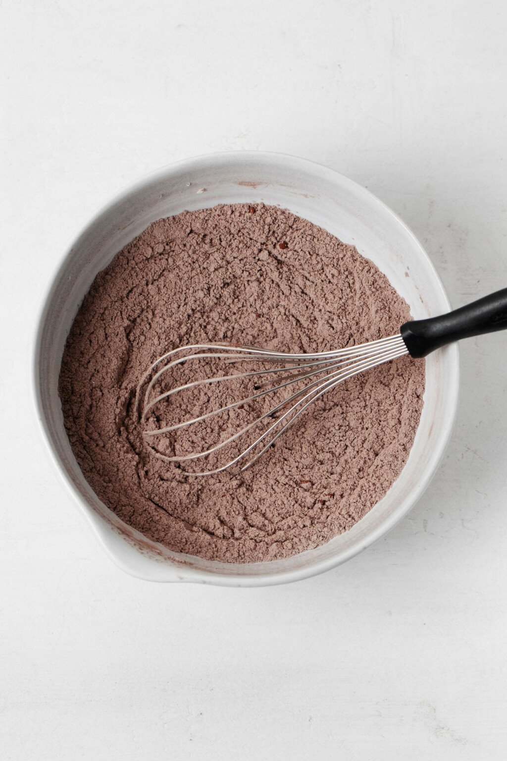 Cocoa powder and flour are being whisked together in a round, white mixing bowl.