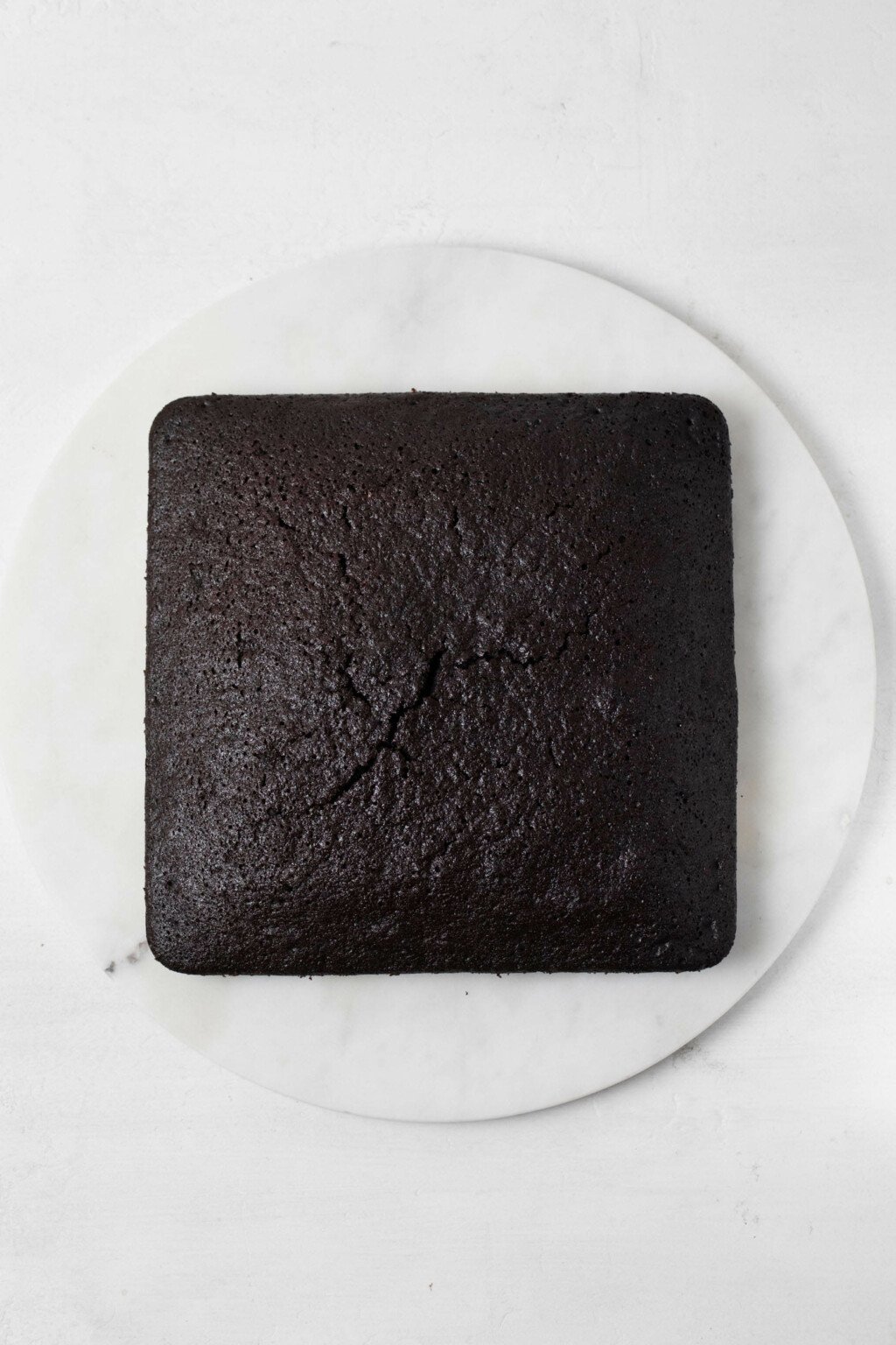 A square, chocolate cake is resting on a round, white marble platter.