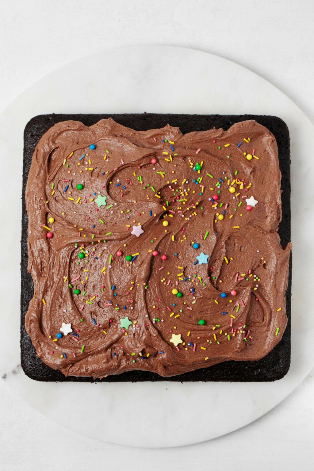 A square vegan chocolate snack cake decorated with buttercream frosting and sprinkles.  It is resting on a white surface.