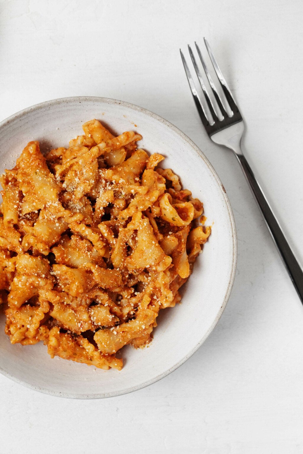 A round, white dish contains a pasta with sun-dried tomato sauce. A silver fork is resting, angled, nearby.