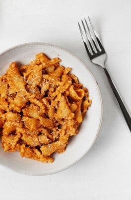 A round, white dish contains a pasta with sun-dried tomato sauce. A silver fork is resting, angled, nearby.