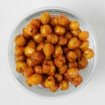 A round, glass mason jar holds crispy roasted chickpeas. It rests on a white surface.