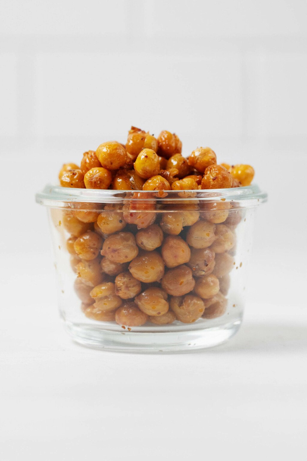 A glass, Weck mason jar is filled with golden, crispy roasted chickpeas. A white tiled surface is visible behind the jar.