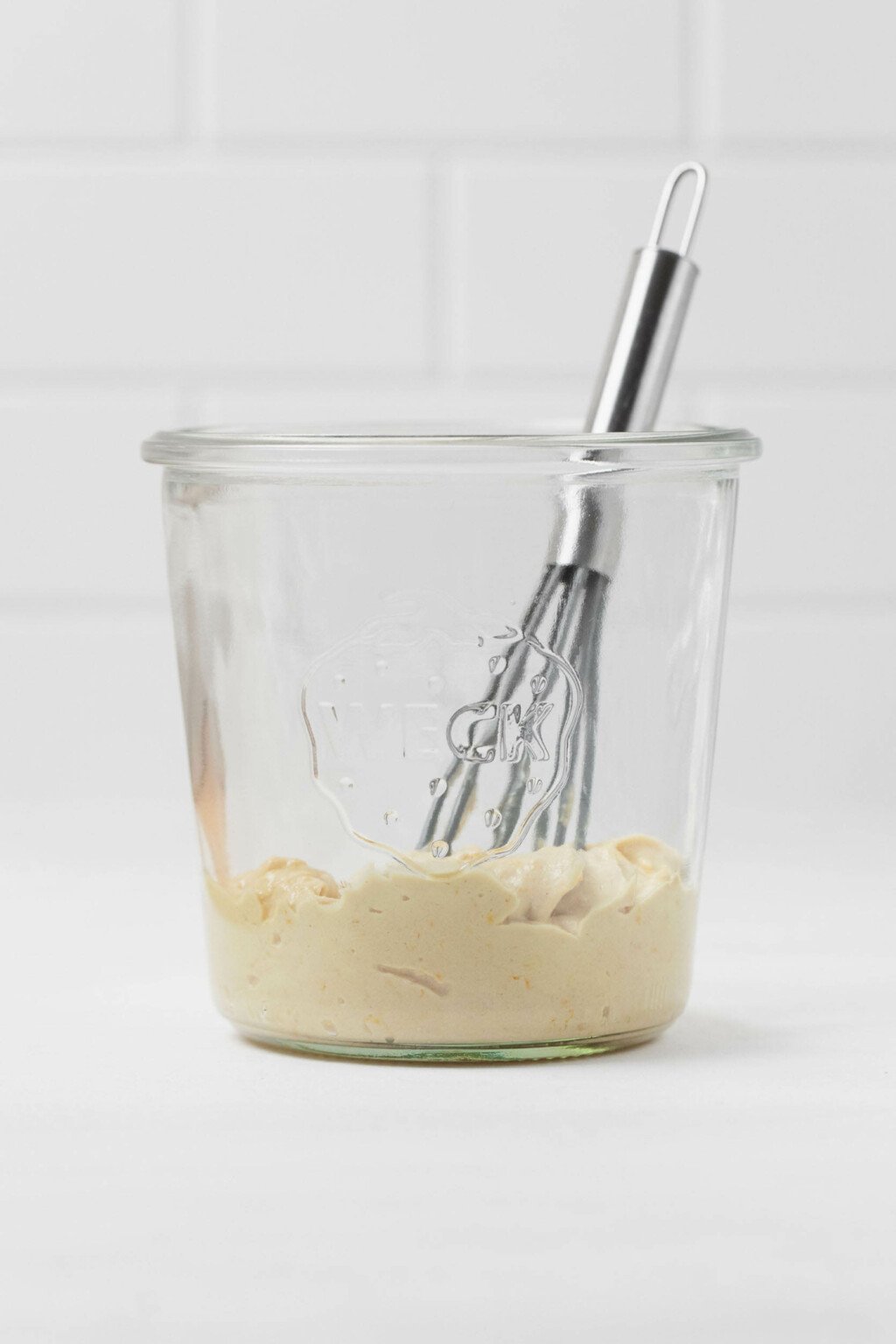 Tahini is in a glass mason jar, along with a small, metal whisk. The jar rests on a white surface.