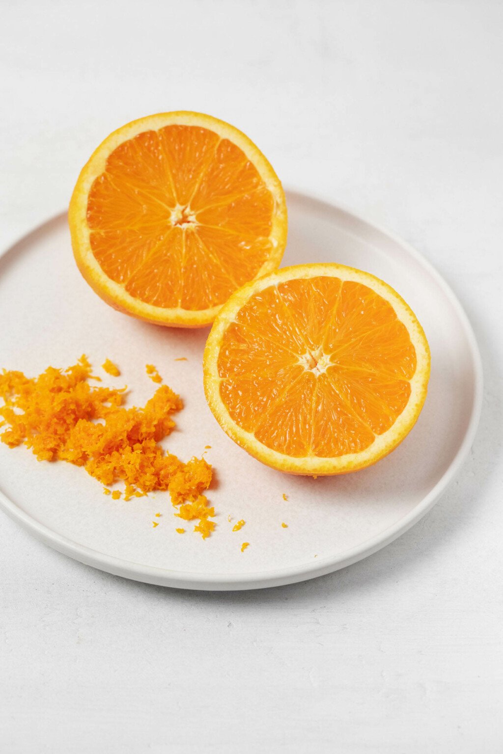 An orange has been sliced in half. It rests on a round white plate, with its zest.