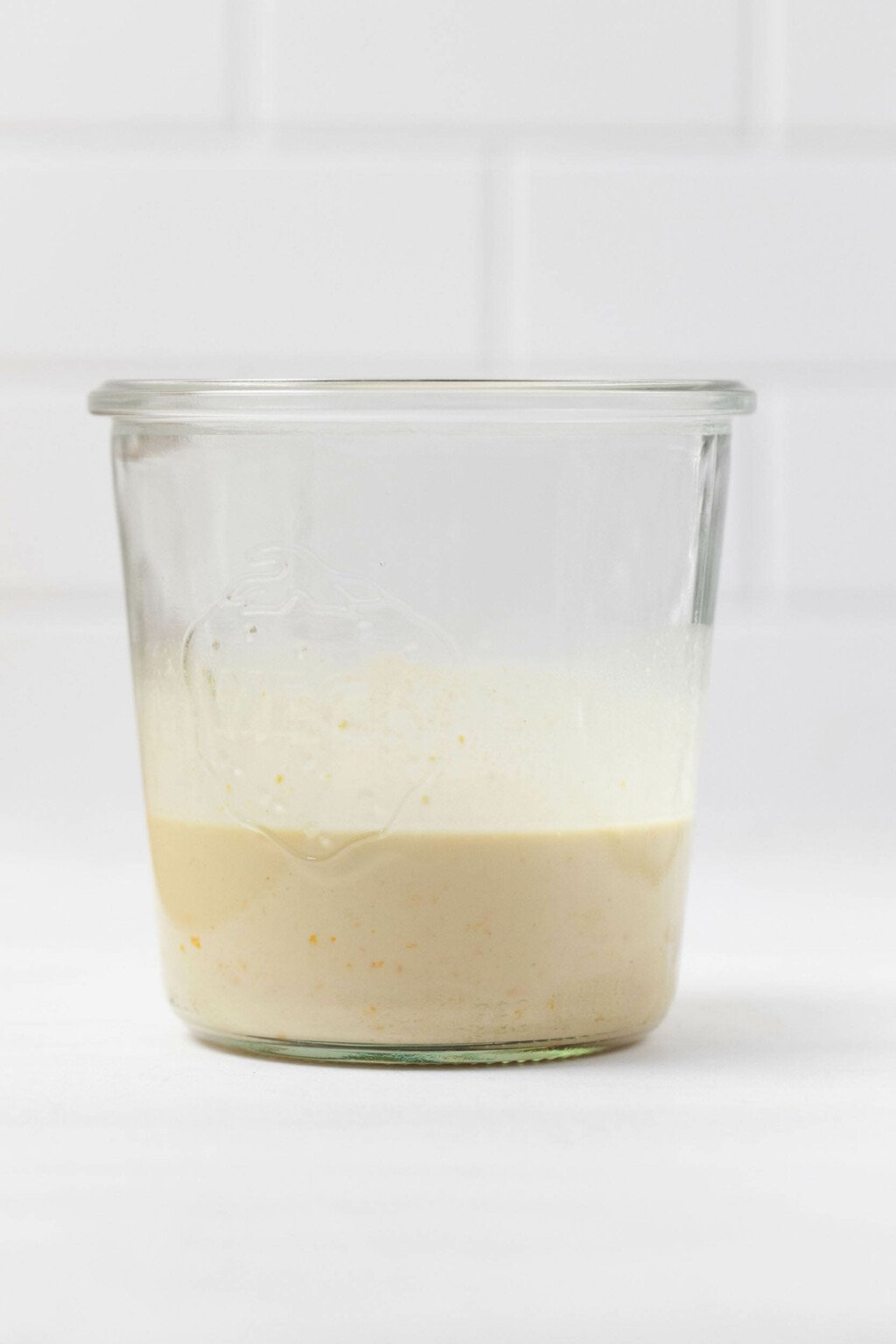 An orange tahini dressing is in a glass, weck mason jar. It rests on a white surface with a tiled backdrop. 