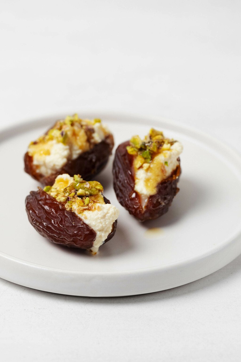 Three dates, which have been stuffed with a plant-based cashew cheese, are resting on a round, white rimmed appetizer plate.