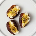 Three sweet and savory stuffed dates, which are topped with pistachio nuts, rest on a white, round appetizer plate.