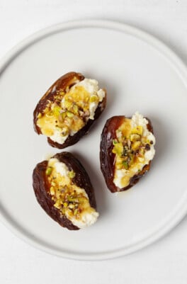 Three sweet and savory stuffed dates, which are topped with pistachio nuts, rest on a white, round appetizer plate.