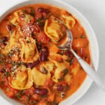 Vegan tortellini soup has been placed into a white bowl. It's being served with a silver spoon.