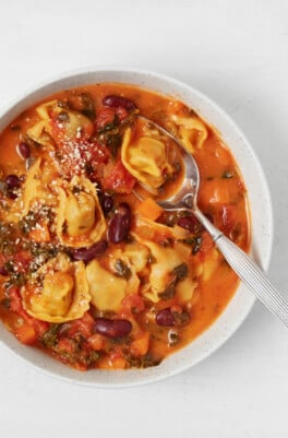 Vegan tortellini soup has been placed into a white bowl. It's being served with a silver spoon.