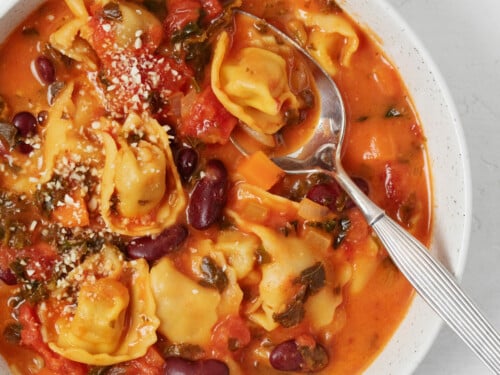 Vegan tortellini soup has been placed into a white bowl. It's being served with a silver spoon.