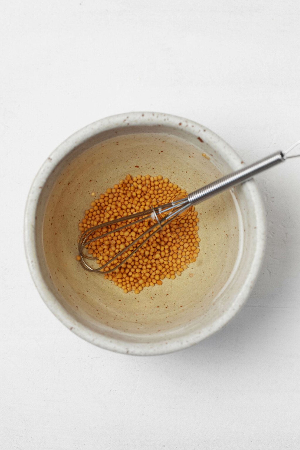 A small, ceramic bowl holds mustard seeds and a mini whisk.