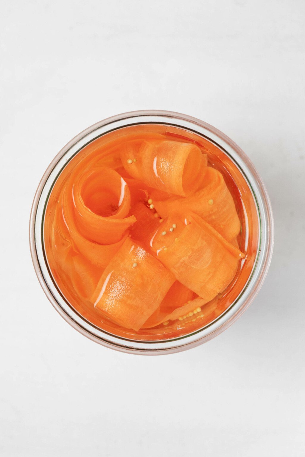 An overhead image of a glass jar containing pickled carrot ribbons. It rests on a white surface.