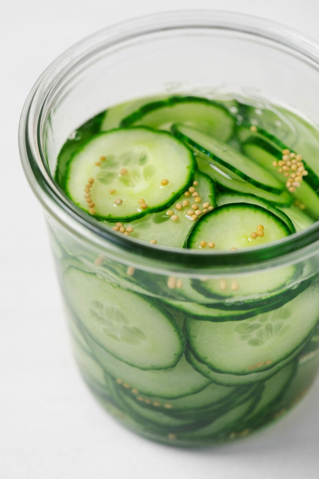 A glass jar is filled with very thinly sliced pickles, liquid, and brown mustard seeds.