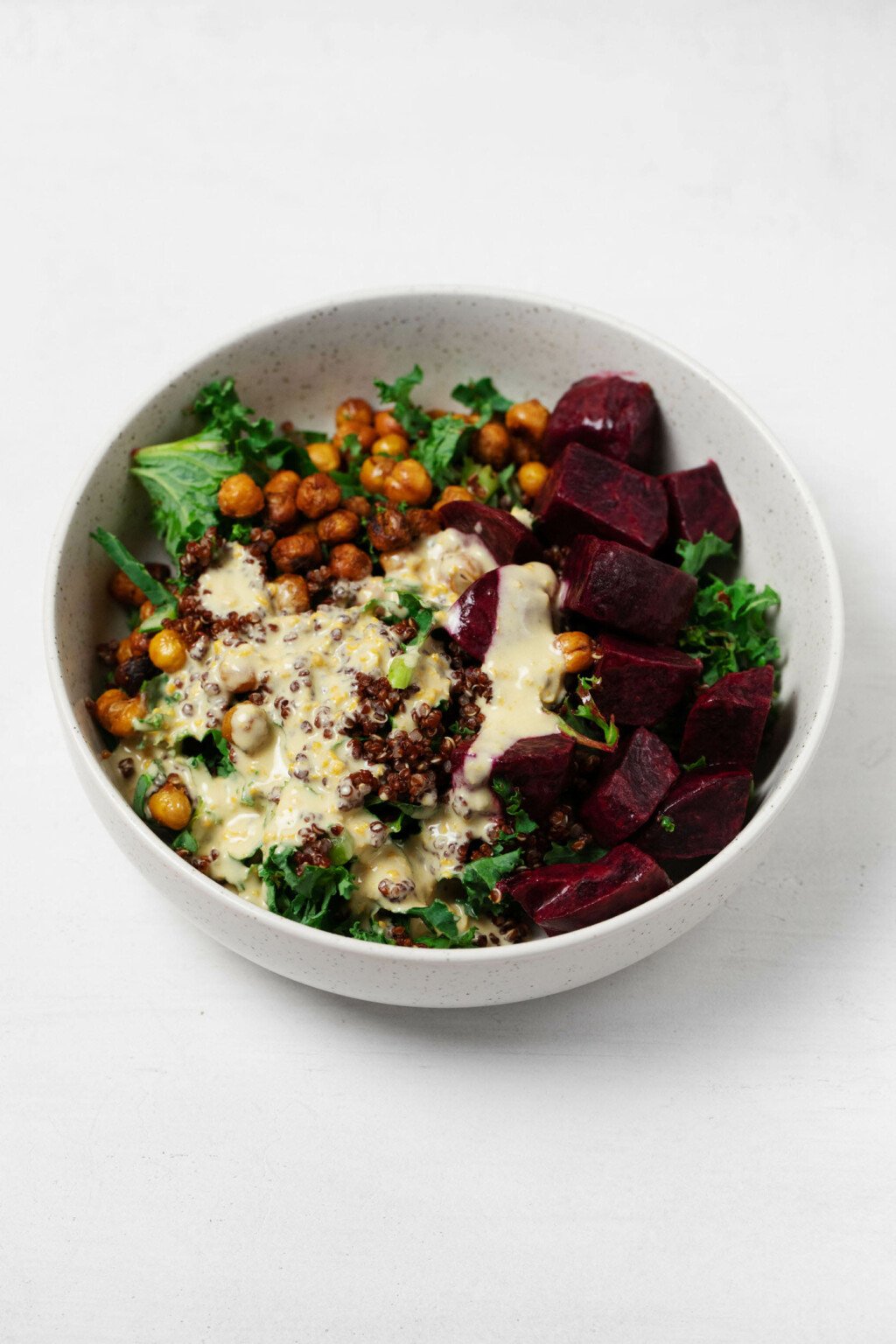 A vegan quinoa beet bowl is resting on a white surface. There's a creamy tahini dressing poured on top of the bowl.