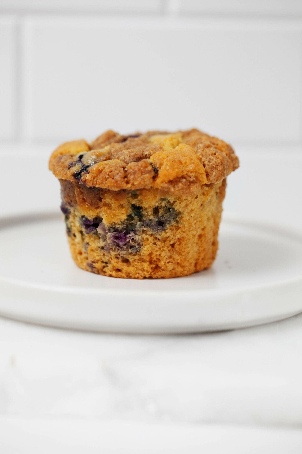 A vegan blueberry crumb muffin rests on a round, rimmed white plate.