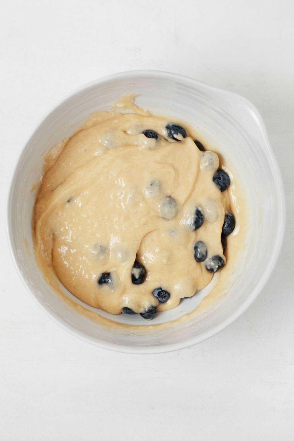 Muffin batter has just had fresh blueberries folded into it, in a white mixing bowl.