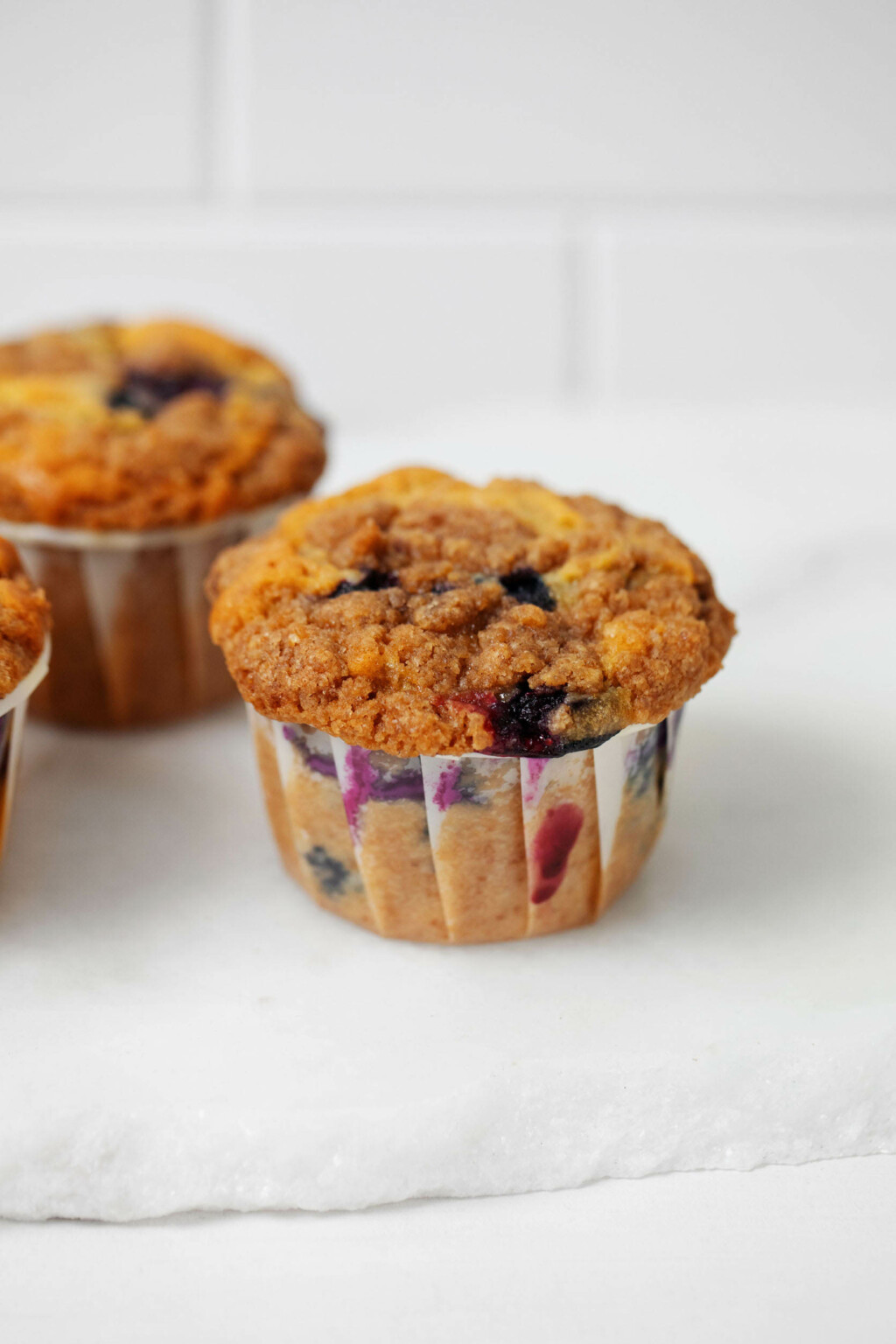 Vegan blueberry crumb muffins are resting on a white surface against a white tile backdrop.