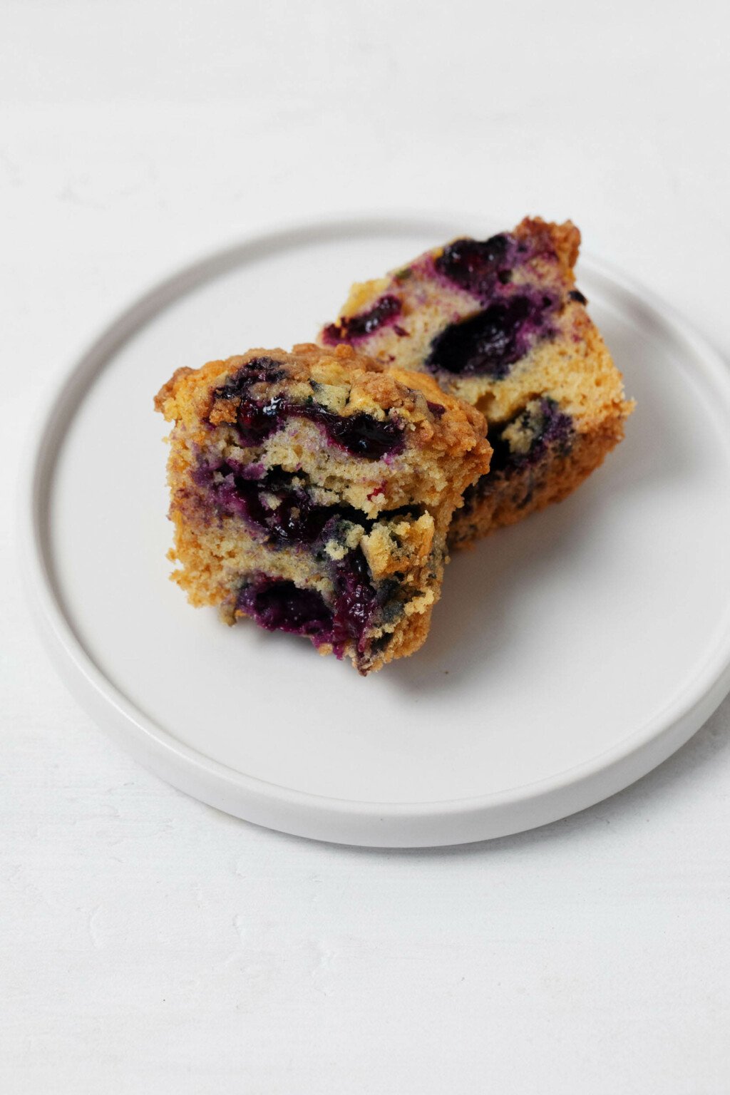 A vegan blueberry crumb muffin is broken apart, resting on a small, round white plate.
