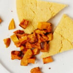 Wedge-shaped slices of a baked, chickpea frittata are served on a round white plate with roasted sweet potato cubes.