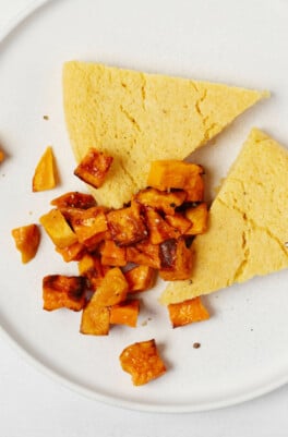 Wedge-shaped slices of a baked, chickpea frittata are served on a round white plate with roasted sweet potato cubes.