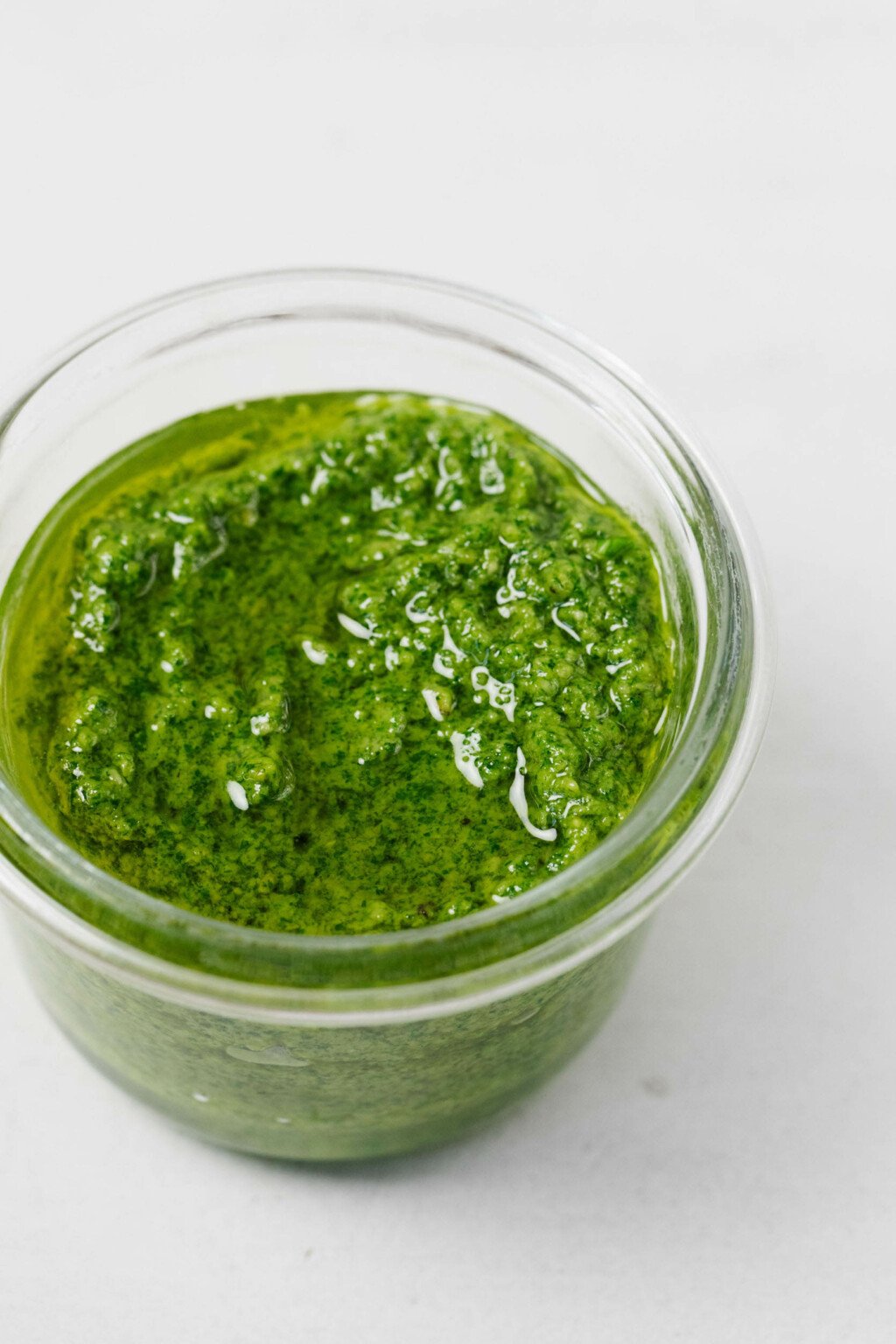 A glass mason jar rests on a white surface. It has been filled with a bright green vegan pesto.