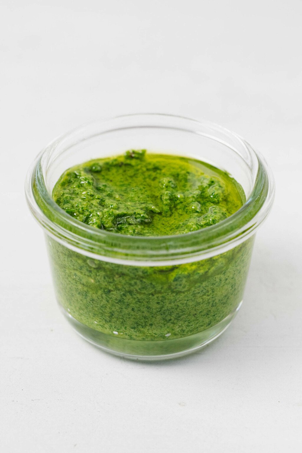 An angled image of a small, clear jar, filled with basil pesto.