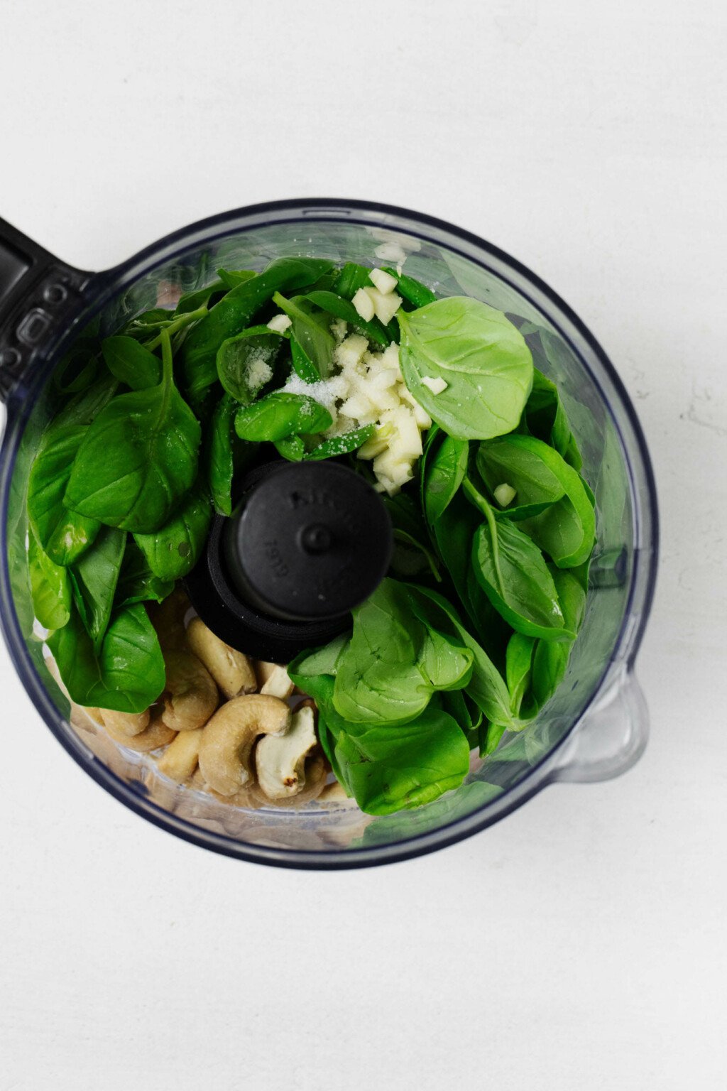 Basil leaves, garlic, and cashews are visible in the bowl of a food processor, pictured overhead. The processor is fitted with an S blade.