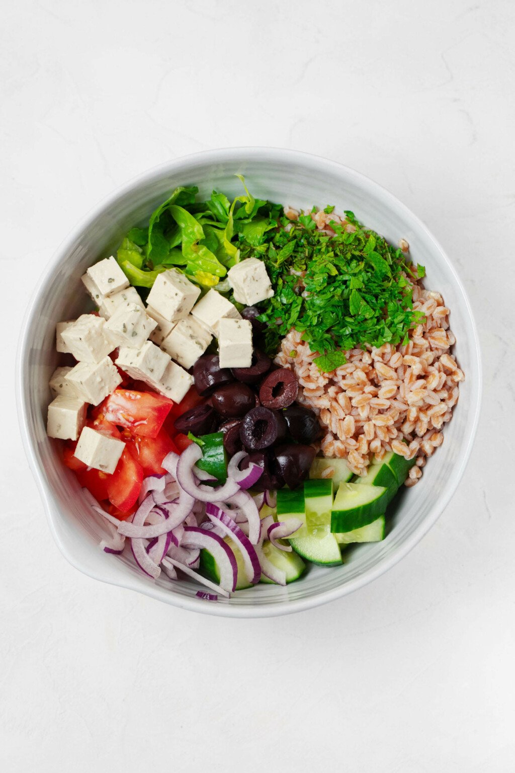 Cubed vegan feta, red onions, cucumber, greens, and barley are resting in a round, white bowl.