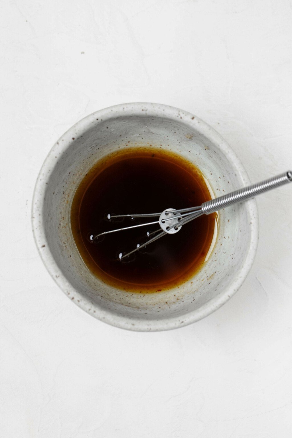 Ingredients for a homemade vinaigrette rest in a small, white pinch bowl with a small whisk.