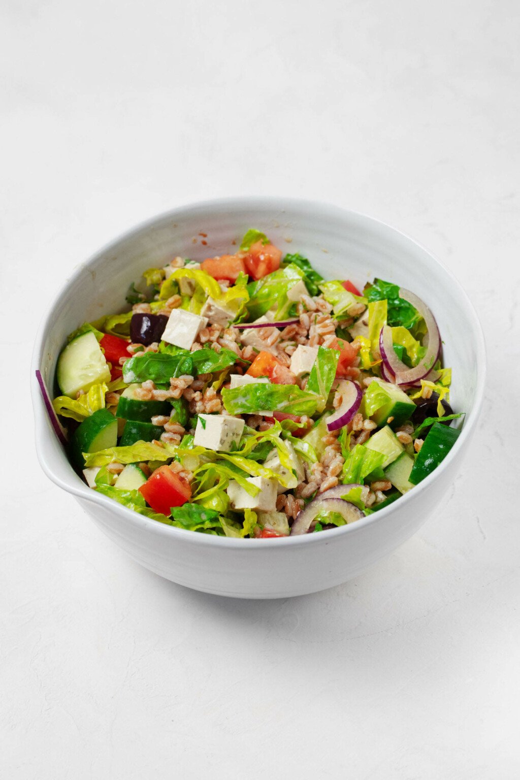 A round, white bowl holds the ingredients for a vegan Greek salad, which is served along with cooked pearled barley.