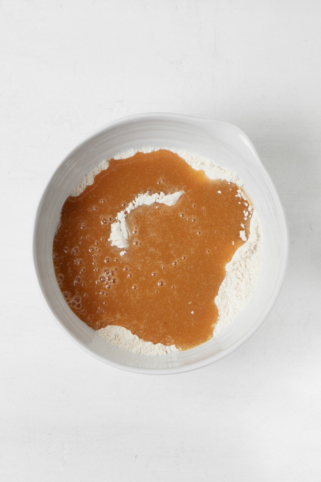 A large mixing bowl holds whisked flour, with a liquid mixture poured into the center. The white mixing bowl rests on a white surface.