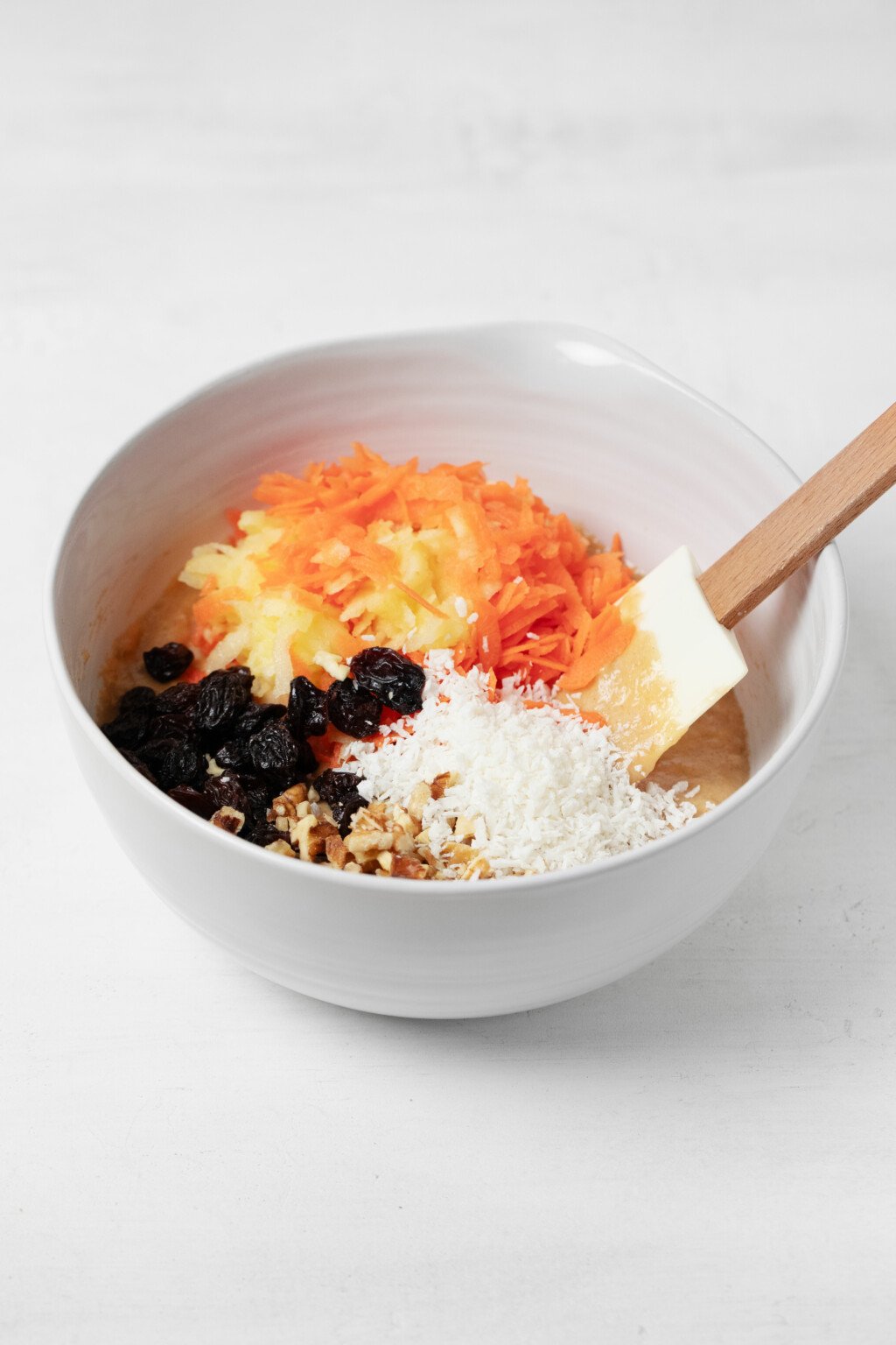 A white mixing bowl is filled with grated carrot, apple, coconut, raisins, and a batter underneath them all.