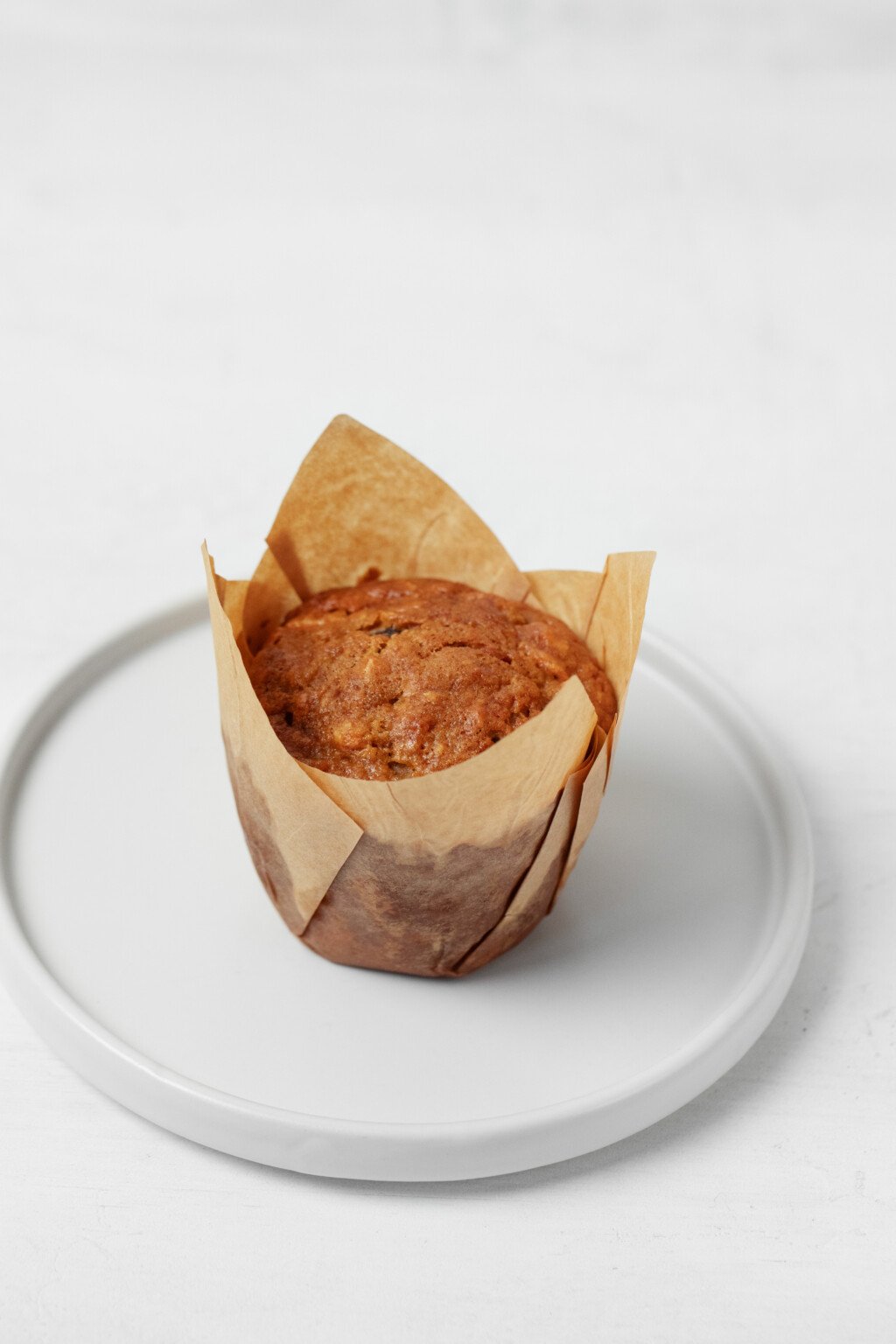 A vegan muffin, which has been wrapped in a brown paper muffin liner, rests on a small, round white plate. 