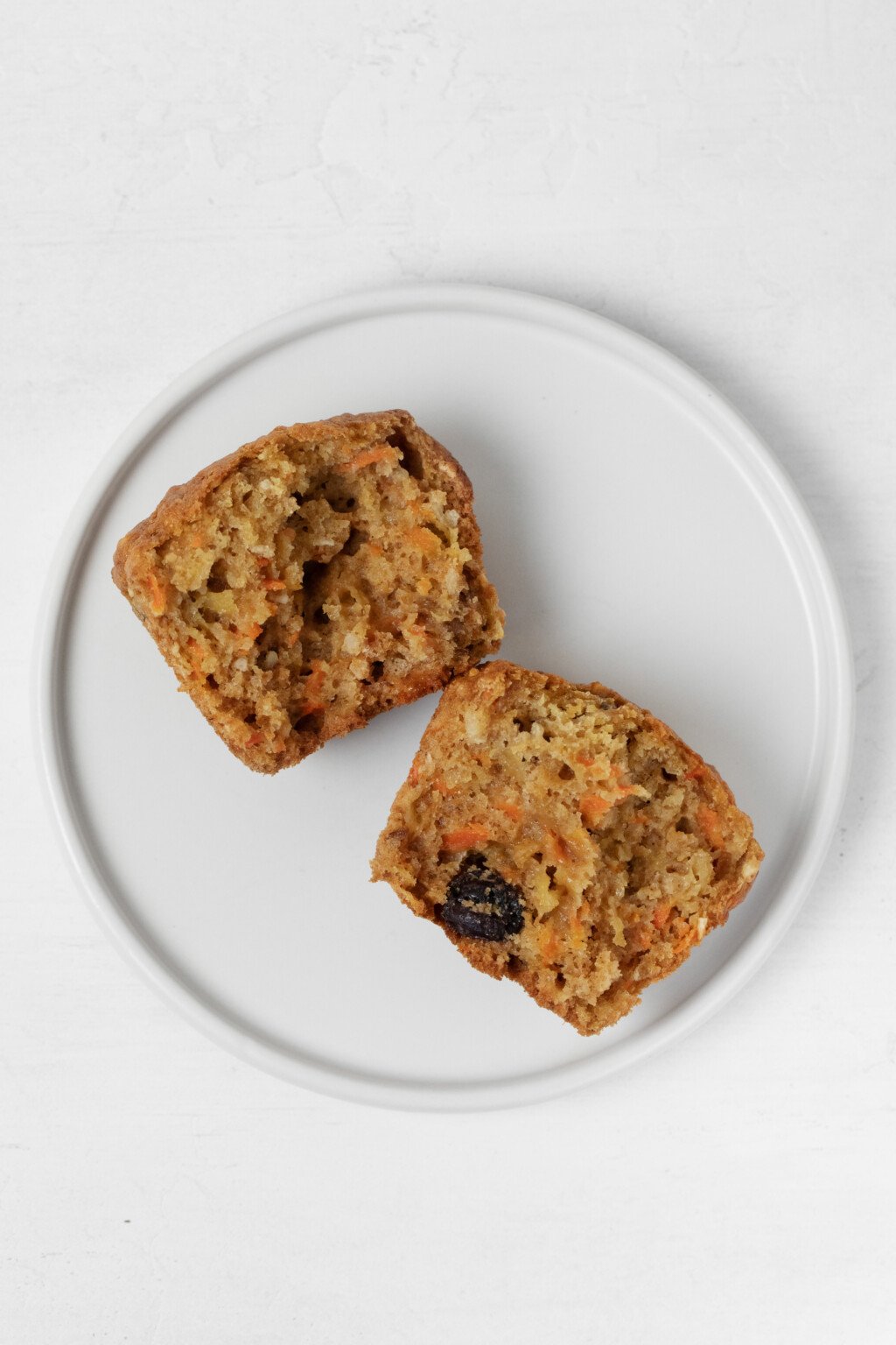 A round, white plate holds a hearty vegan morning glory muffin, which has been broken open to reveal a soft interior.