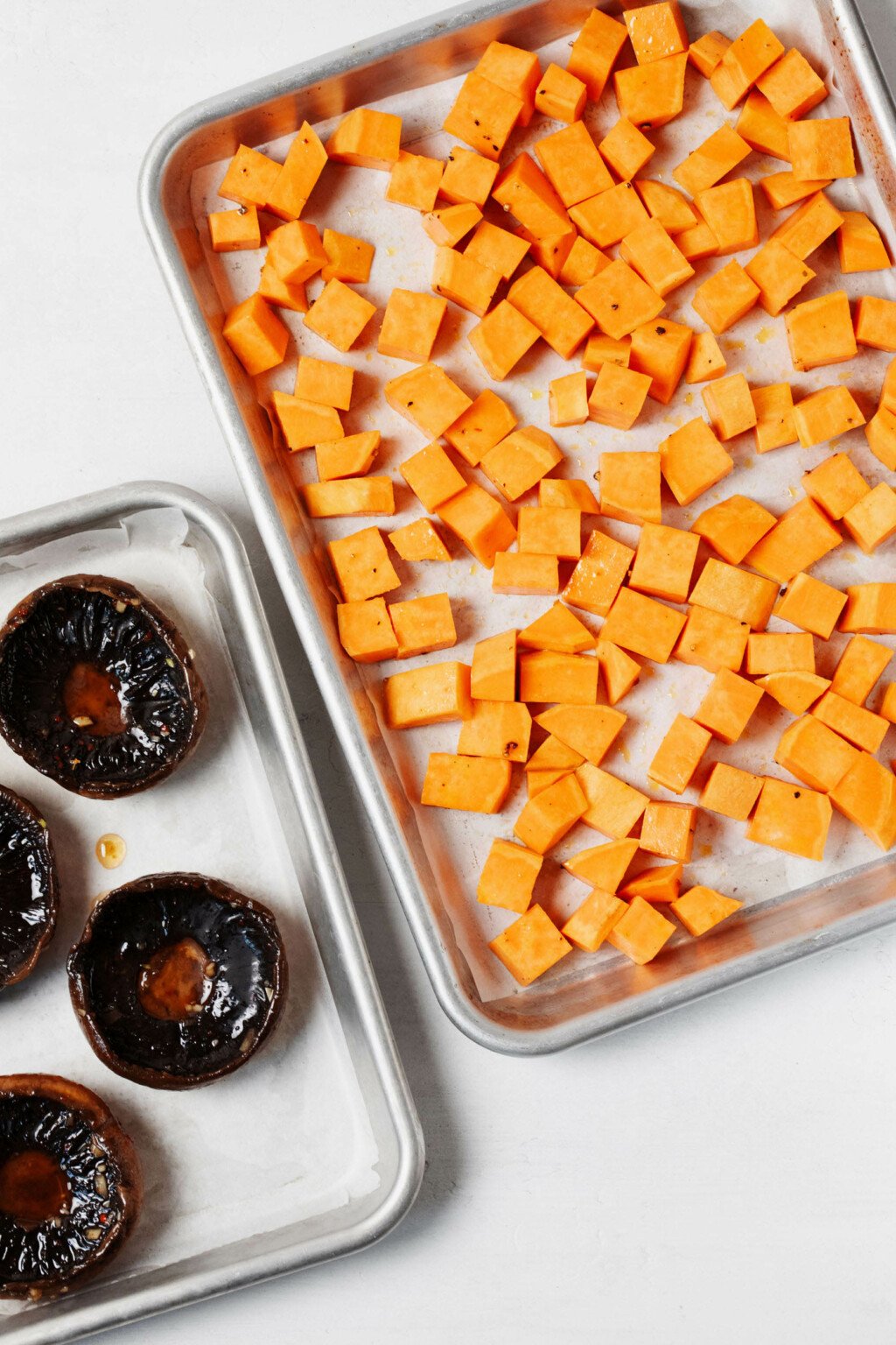 Two silver baking sheets hold portobello caps and cubed sweet potatoes for roasting.
